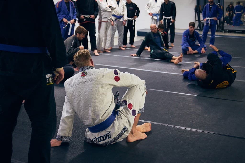 A Jiu Jitsu class on the mats - a seated practitioner in the foreground with the room training behind him